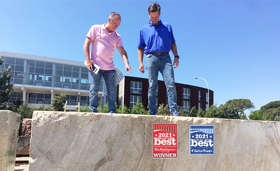 A man in a pink shirt and jeans points down at a slab while a man in a blue shirt and jeans looks.